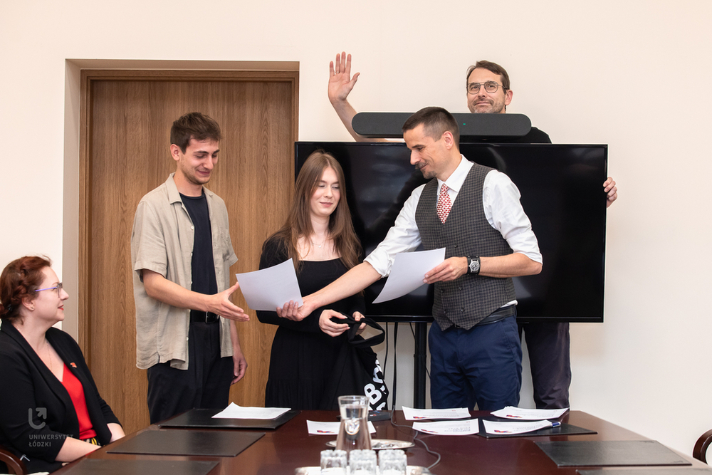Moment wręczania dyplomów od prof. Krzysztofa Pabisa. Na drugim planie Rektor, prof. Rafał Matera machający do fotografa zza dużego ekranu telewizora.