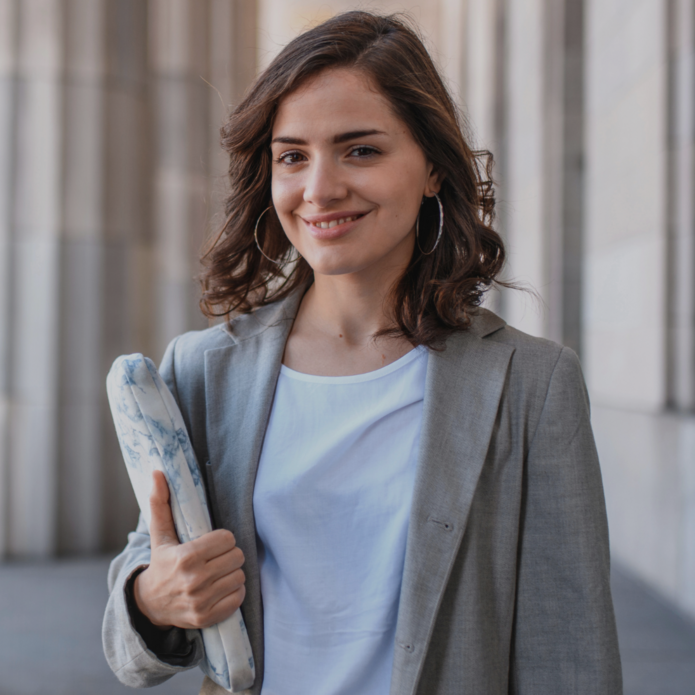 Młoda kobieta o brązowych, falowanych włosach i delikatnym uśmiechu stoi na tle monumentalnych, kamiennych kolumn. Ma na sobie szarą marynarkę i białą bluzkę, w ręku trzyma zwinięty materiał lub etui w marmurkowy wzór, pokazując kciuk uniesiony w geście aprobaty. Nosi duże, okrągłe kolczyki i sprawia wrażenie pewnej siebie oraz profesjonalnej.